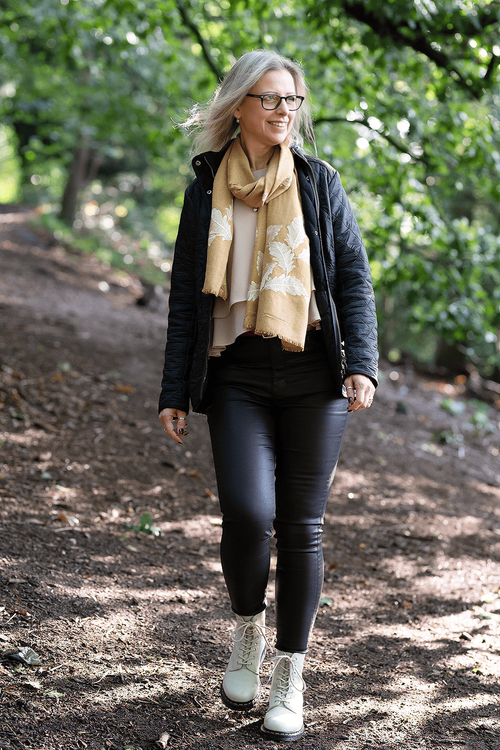 Author Michelle Townsend smiling as she walks forward along a dappled woodland path, wearing a yellow patterned scarf and white boots.