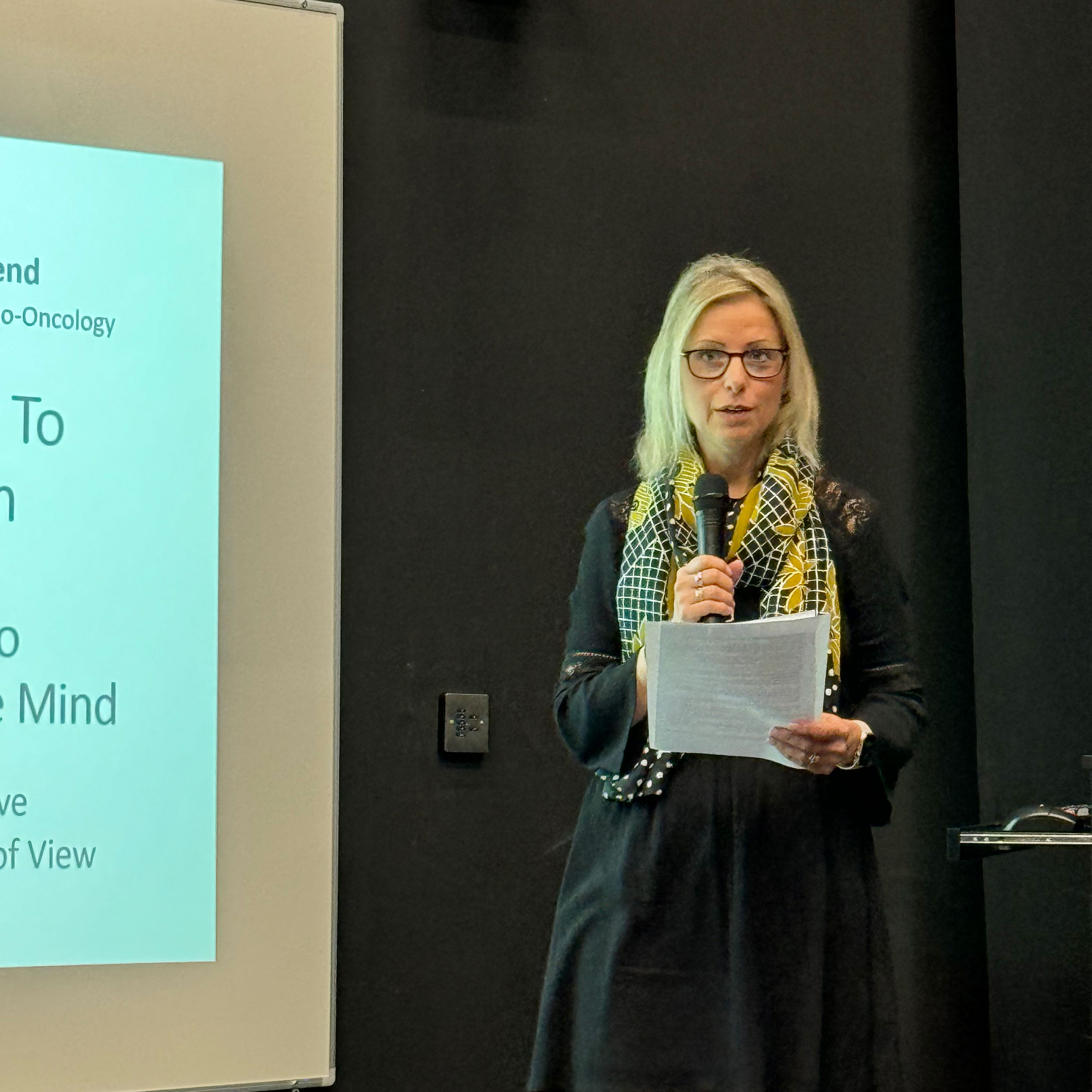 Michelle Townsend holding notes and speaking into a microphone at the BCU European Cancer Conference. She is wearing a black dress and a patterned scarf, standing next to a partially visible presentation screen about understanding the mind.
