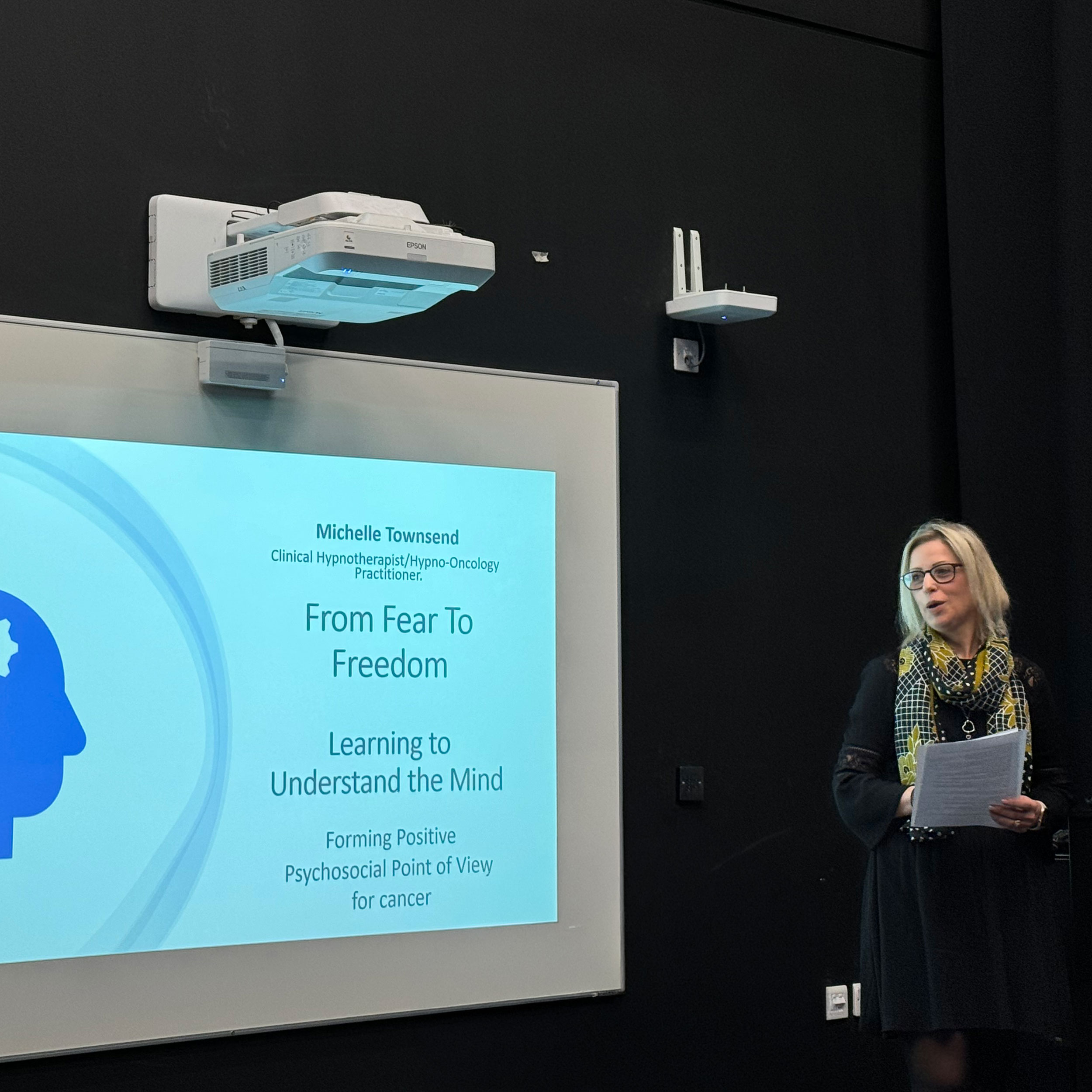 Clinical Hypnotherapist Michelle Townsend holding notes and speaking at the BCU European Cancer Conference. She is standing next to a large projection screen displaying her presentation titled 'From Fear To Freedom: Learning to Understand the Mind', which discusses forming a positive psychosocial point of view for cancer.