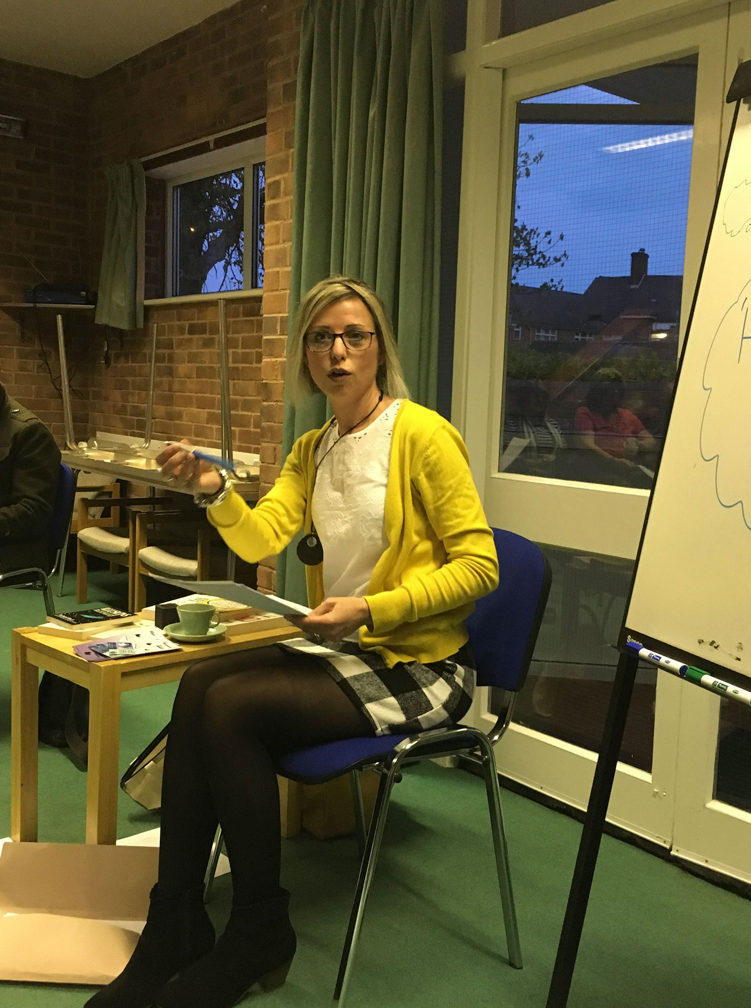A woman with blonde hair and glasses sits on a blue chair, speaking to an unseen audience while gesturing with a blue pen and holding notes. She is wearing a yellow cardigan, a white top, a black-and-white checkered skirt, dark tights, and black ankle boots. To her left is a small wooden table holding books, papers, and a light green teacup. A whiteboard is partially visible on the right side of the frame.