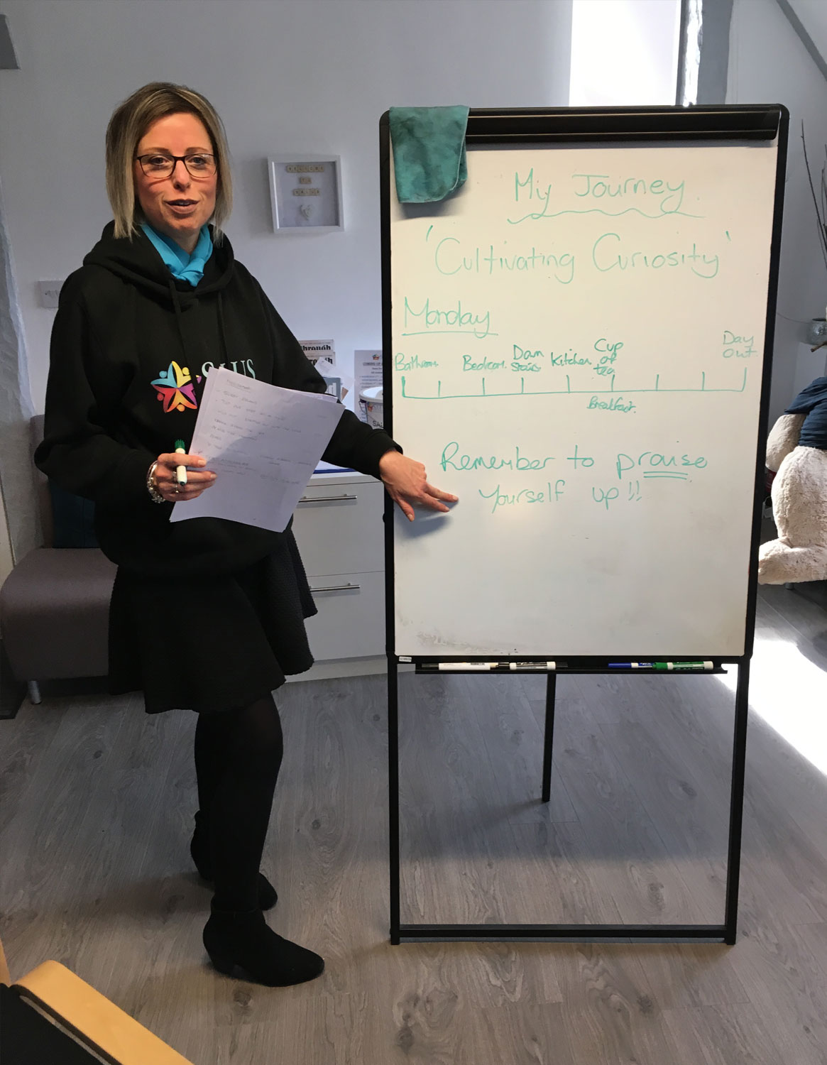 Here is an alt tag for the image: "A woman in a black logo hoodie and skirt standing next to a whiteboard titled 'My Journey 'Cultivating Curiosity''. Holding notes in one hand, she points to the phrase 'Remember to praise Yourself up!!' written at the bottom of the board. The whiteboard also displays a handwritten timeline mapping out daily activities for 'Monday'.