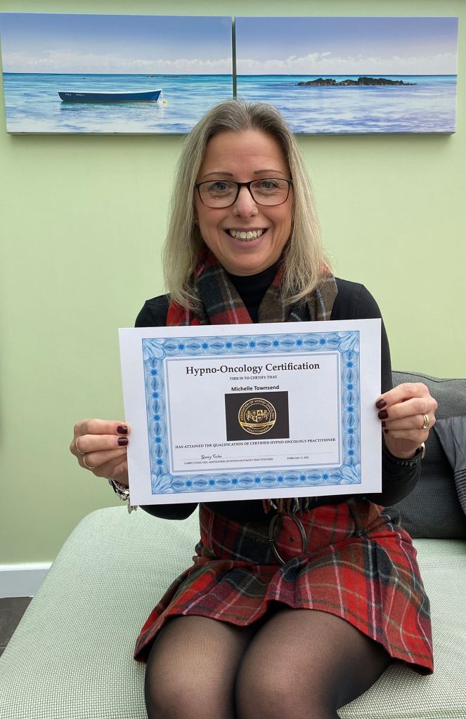 A smiling woman, Michelle Townsend, proudly holds up her "Hypno-Oncology Certification," which states she has attained the qualification of Certified Hypno-Oncology Practitioner. She is sitting on a light green sofa, wearing glasses, a black long-sleeved top, and a matching red plaid scarf and skirt.