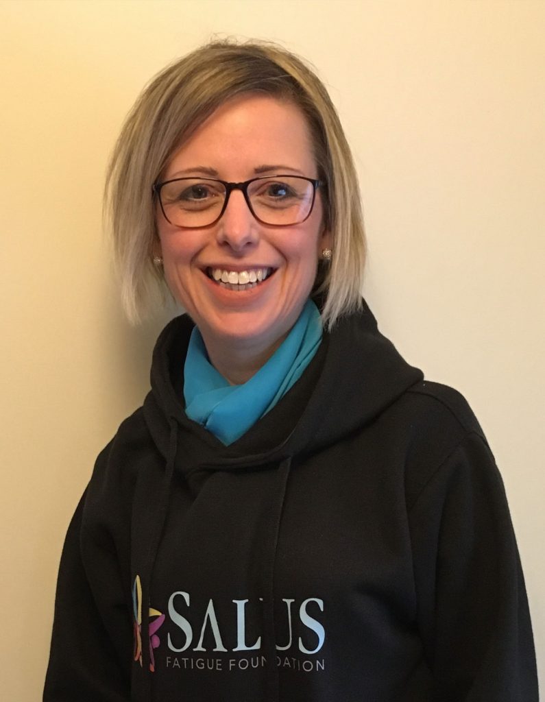 A headshot of a smiling woman with short blonde hair and glasses. She is wearing a bright blue top under a black hoodie featuring the 'SALUS FATIGUE FOUNDATION' logo, standing against a plain light-coloured background.