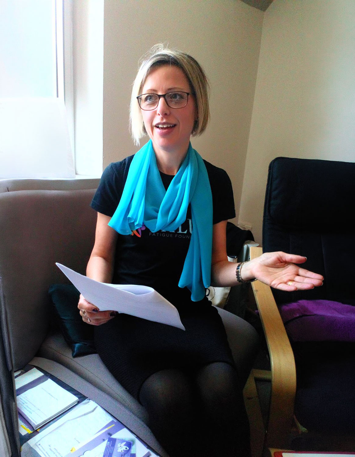 A woman with short blonde hair and glasses seated in a grey chair near a window, speaking and gesturing outward with an open hand. She is holding papers in her lap and wearing a black outfit with a bright blue scarf and a partial 'Fatigue Foundation' logo. Several brochures and papers rest on the chair cushion beside her.