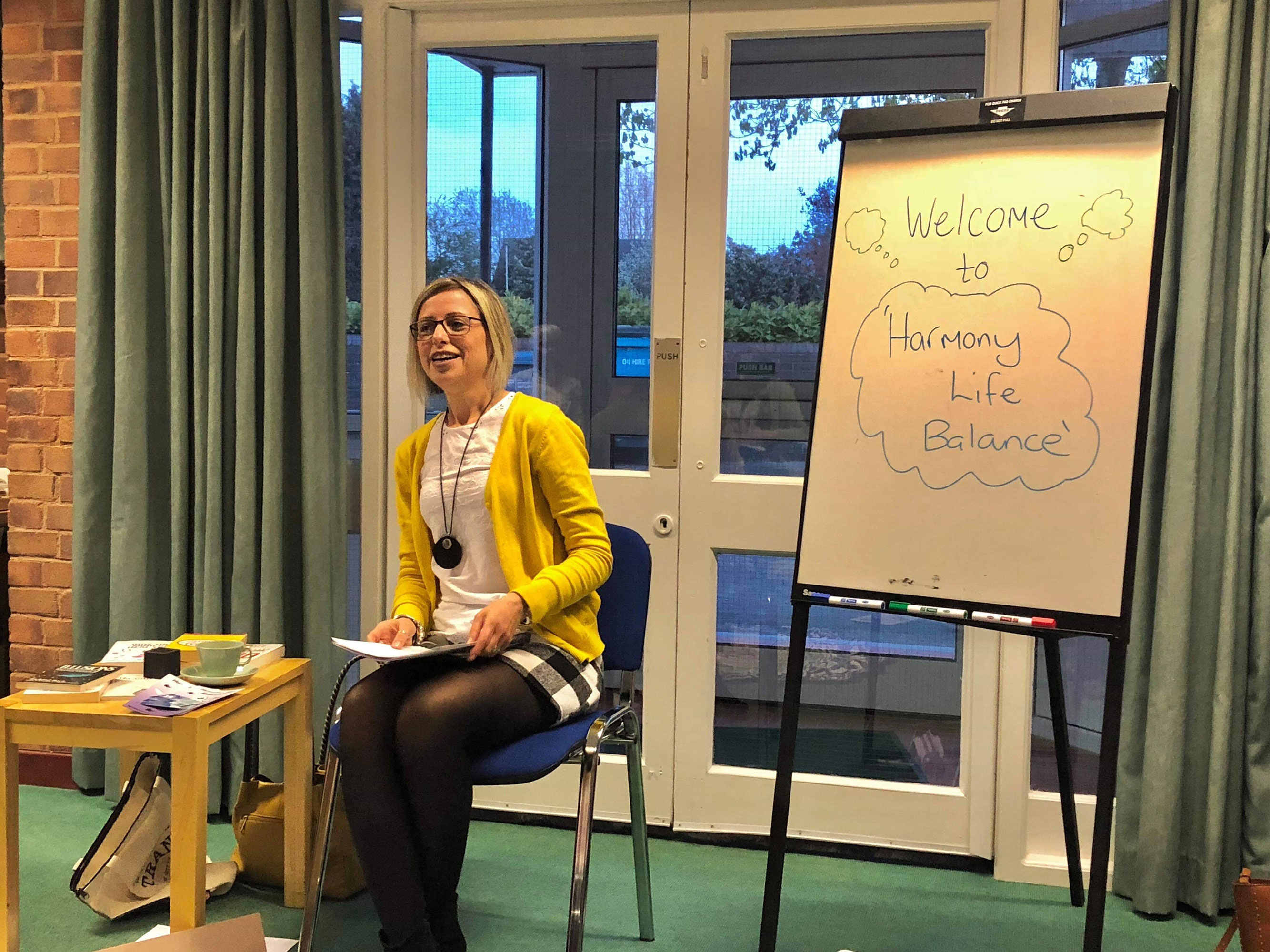 Here is an alt tag for the second image: Alt text: A woman with blonde hair and glasses sits smiling on a blue chair, holding notes in her lap. She is wearing a yellow cardigan, a white top with a long black pendant necklace, a black-and-white checkered skirt, and dark tights. To her left is a small wooden table holding books, papers, and a light green teacup. Behind her are white, glass-paned double doors looking outside. To her right stands a whiteboard on an easel with the handwritten text "Welcome to 'Harmony Life Balance'" inside a drawn cloud.