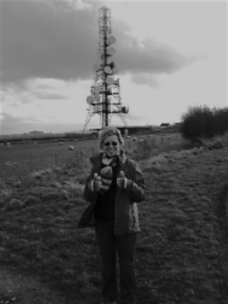 A grainy, black-and-white photograph of a smiling woman with short hair and glasses, wearing a warm coat and gloves. She is standing in a grassy field and giving a double thumbs-up directly to the camera. Behind her, dominating the skyline under a cloudy sky, is a tall, intricate telecommunications tower heavily loaded with antennas.