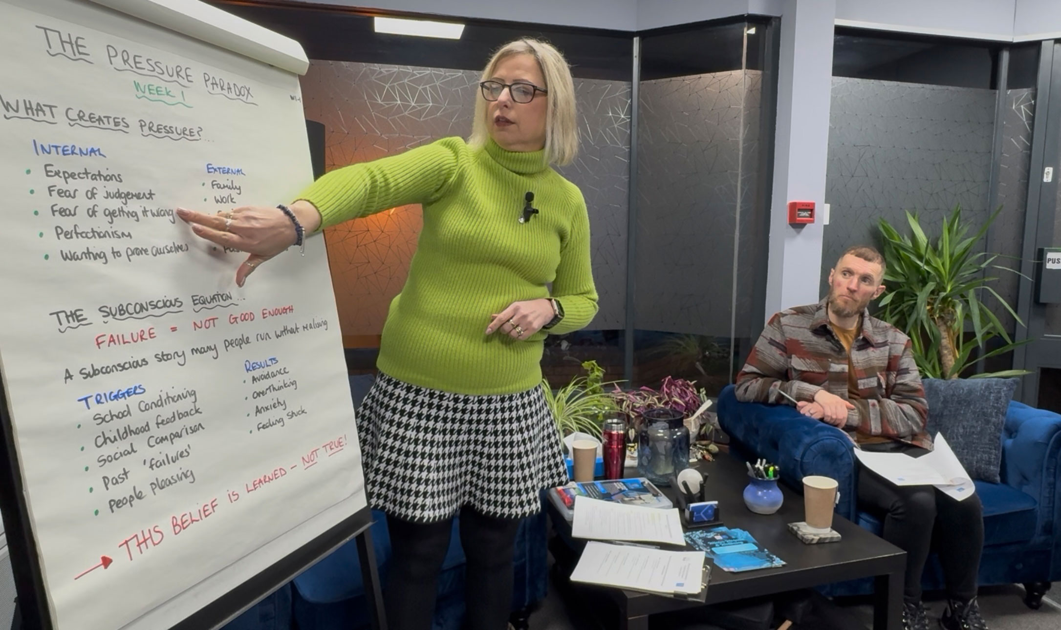 Michelle standing and speaking to an audience, wearing a green turtleneck and patterned skirt. She is standing next to a flip chart with handwritten notes about emotional clarity. David sits beside her on a blue velvet sofa, holding notes and listening closely.