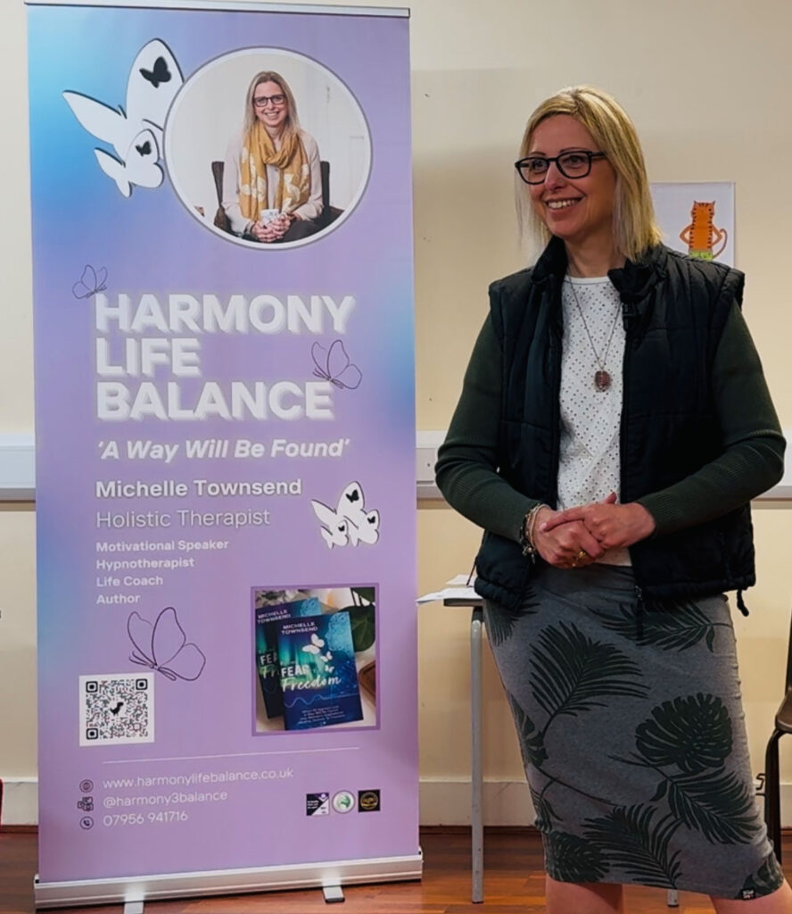 Michelle standing and smiling brightly next to her purple 'Harmony Life Balance' pull-up banner. She is wearing a black vest, an olive top, and a leaf-patterned skirt. The banner details her work as a Holistic Therapist and Author, featuring a photo of her book 'From Fear to Freedom'.