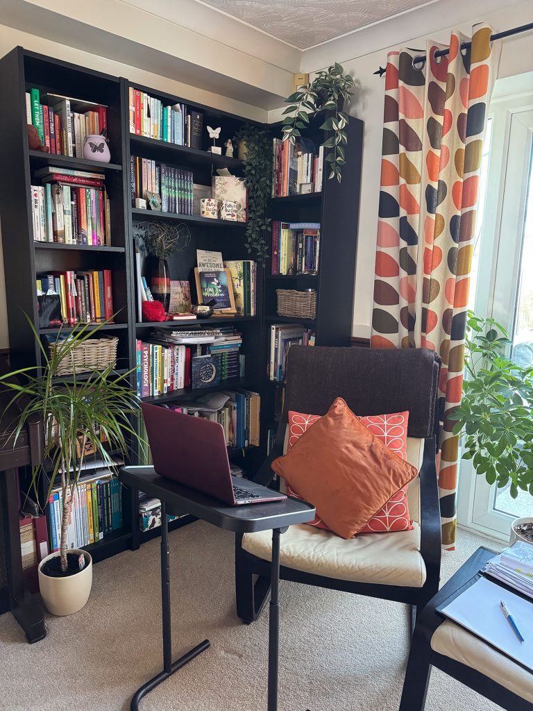 A cosy home therapy space featuring a cream armchair with orange cushions facing a maroon laptop on a small adjustable table. The room is decorated with tall black bookshelves filled with books, various houseplants, and retro-patterned curtains by a bright window.