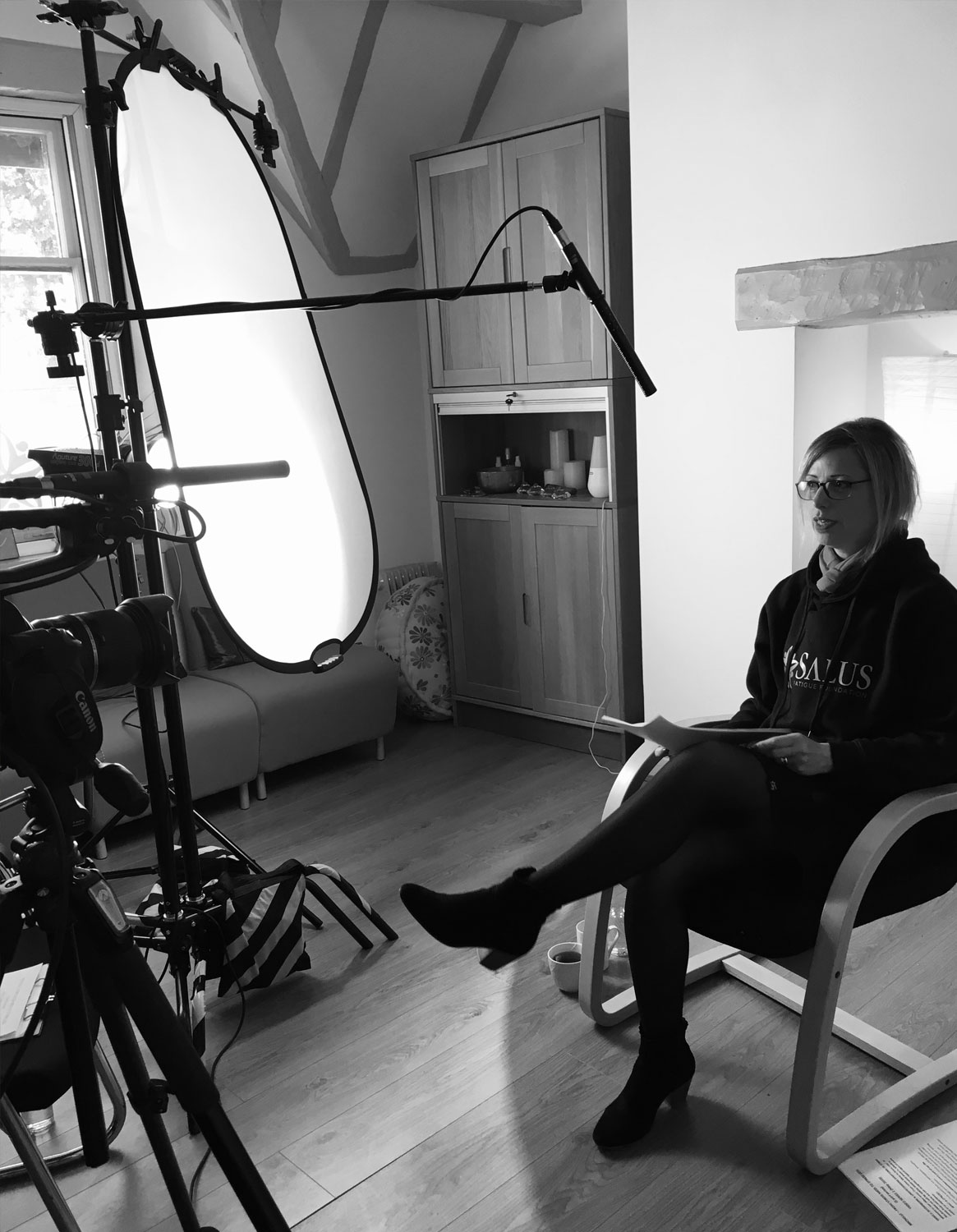 A black and white behind-the-scenes photograph of a video shoot. A woman wearing glasses and a dark hoodie with the 'SALUS FATIGUE FOUNDATION' logo sits cross-legged in a light wooden chair, holding a document. She is positioned in front of a camera on a tripod, a large illuminated light diffuser, and an overhead boom microphone.