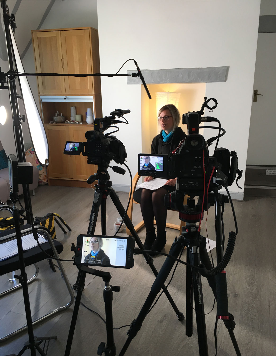 Here is an alt tag for the image: "A behind-the-scenes view of a professional video shoot. A woman with short blonde hair and glasses sits in a chair holding papers, being filmed by a multi-camera setup. The room is filled with video production equipment, including several cameras mounted on tripods, an external field monitor, a large lighting diffuser, and an overhead boom microphone.